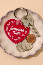 Keychain with 'feminine rage' on a heart-shaped tag on a beige background