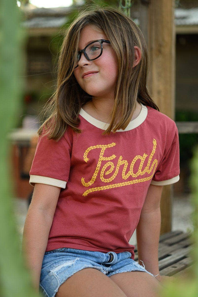 Young girl wearing a red shirt with yellow text, sitting outdoors among plants.