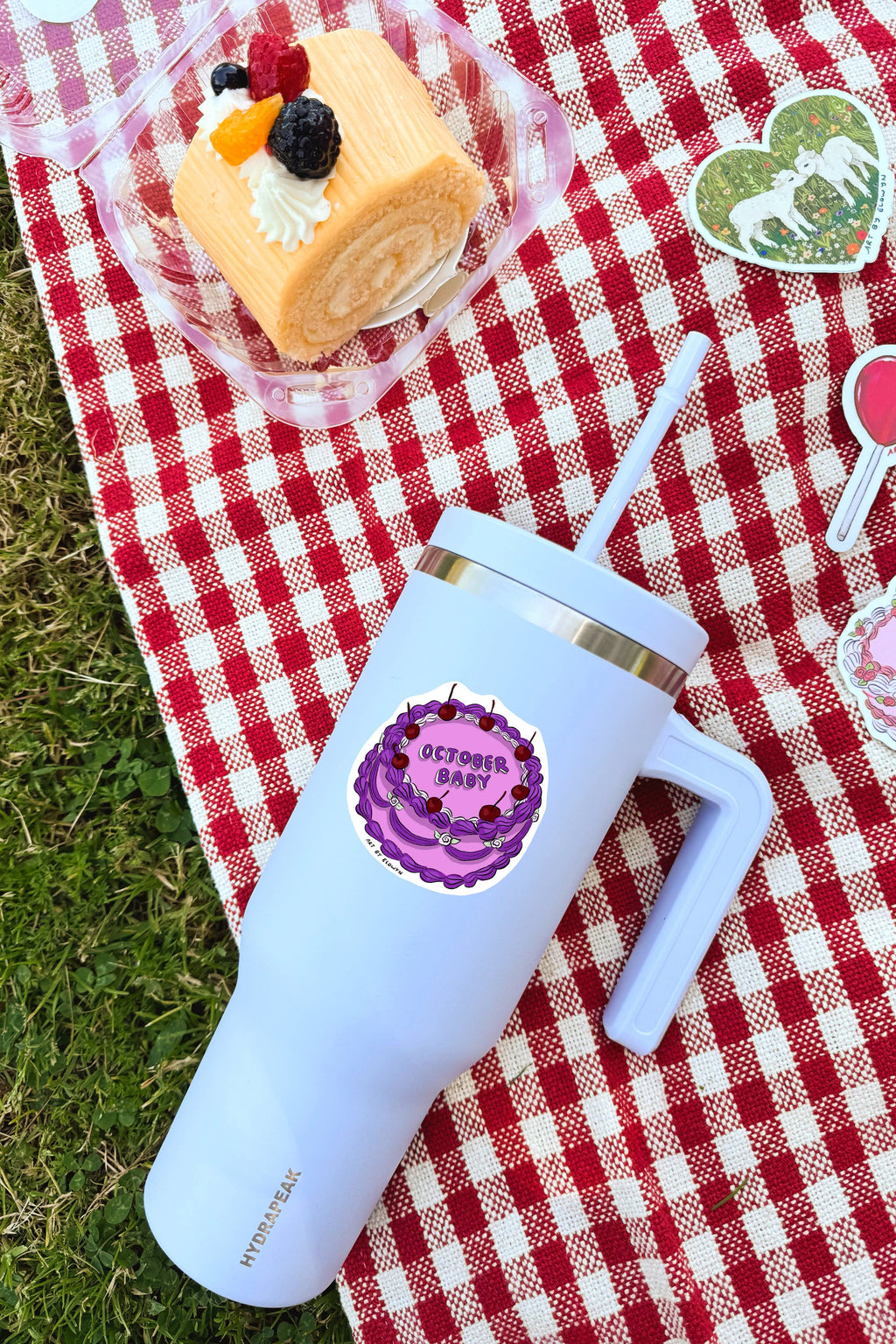 White tumbler with purple lid on a red and white checkered tablecloth with cake and decorations.