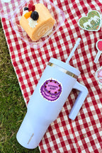 White tumbler with purple lid on a red and white checkered tablecloth with cake and decorations.