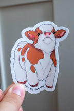 Hand holding a sticker of a cow with orange spots against a neutral background