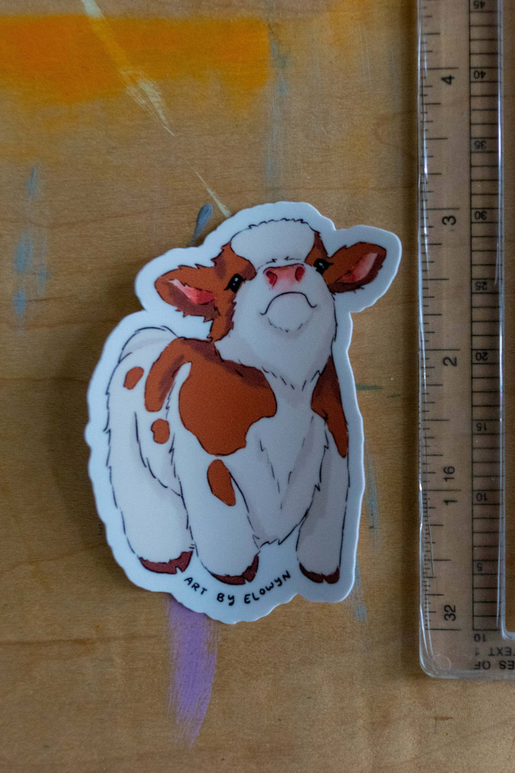 Sticker of a cow with orange spots next to a ruler on a wooden surface