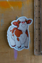 Sticker of a cow with orange spots next to a ruler on a wooden surface