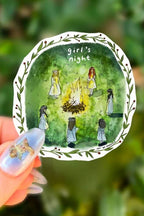 Decorative plate with illustration of 'girl's night' and people around a campfire, held against a green leafy background.