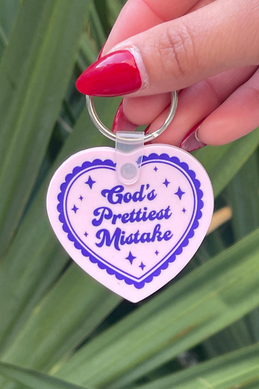 Heart-shaped keychain with 'God's Prettiest Mistake' text held by a hand against a green leafy background
