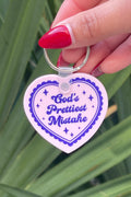 Heart-shaped keychain with 'God's Prettiest Mistake' text held by a hand against a green leafy background