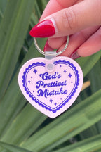 Heart-shaped keychain with 'God's Prettiest Mistake' text held by a hand against a green leafy background
