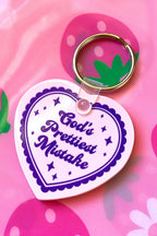 Heart-shaped keychain with 'God's Prettiest Mistake' text on a pink background with strawberry and flower designs.