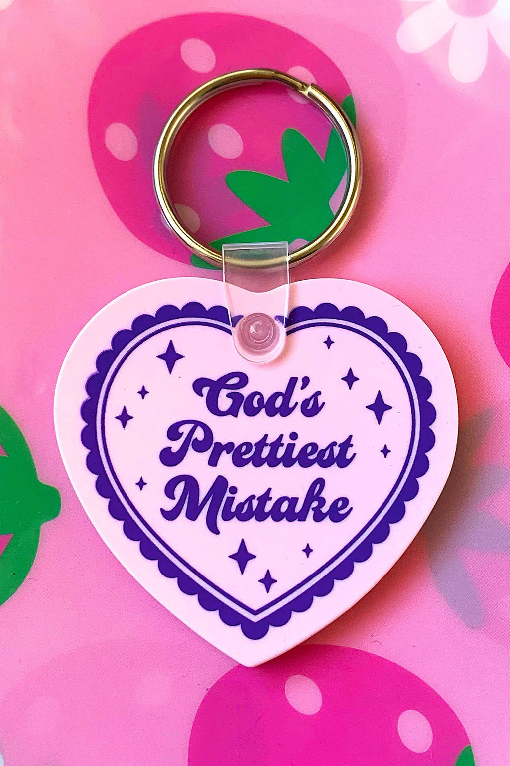 Heart-shaped keychain with 'God's Prettiest Mistake' text on a pink background with strawberry and flower designs.