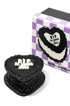 Black heart cake-shaped candle with white text next to a purple and white checkered box. rip to your 20's