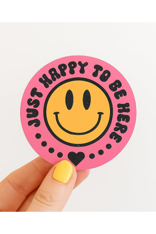 Sticker with smiley face and text 'Just happy to be here' held by a hand with yellow nail polish on a white background.