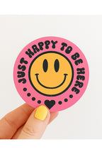 Sticker with smiley face and text 'Just happy to be here' held by a hand with yellow nail polish on a white background.