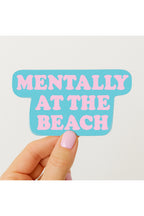 Hand holding a sticker with 'Mentally at the Beach' text on a light background