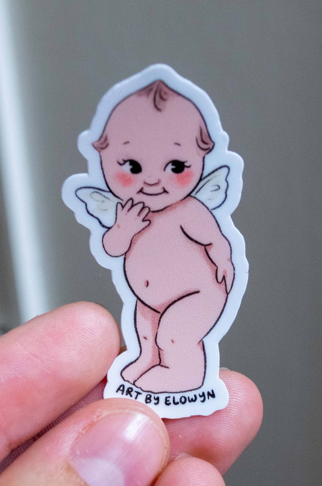 Hand holding a small sticker of a cartoon baby with angel wings against a neutral background