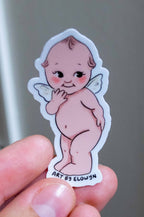 Hand holding a small sticker of a cartoon baby with angel wings against a neutral background