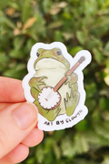 Hand holding a sticker of a frog playing a banjo with a green leafy background