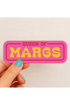 Pink and yellow 'Queen of Margs' sticker held by a hand with teal nail polish on a beige background