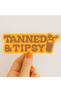 Hand holding a 'Tanned & Tipsy' sticker with a tropical drink graphic on a light background