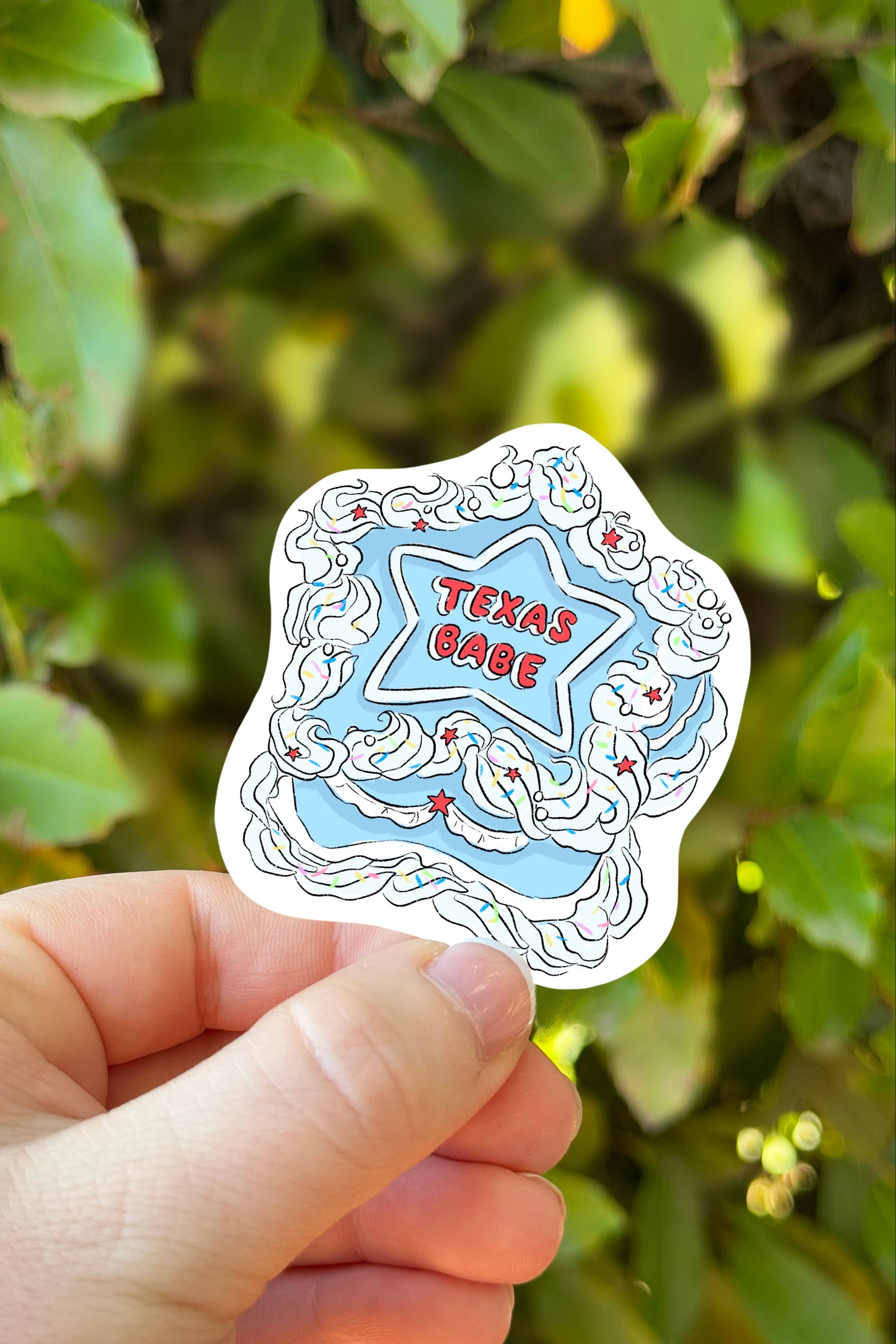 Hand holding a decorative sticker with 'Texas Cake' text against a green leafy background