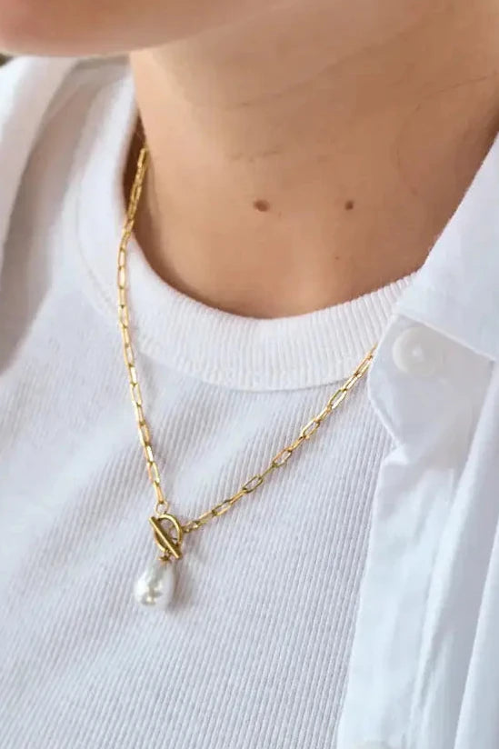 Person wearing a gold necklace with a clear pendant on a white shirt.