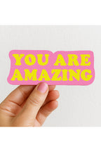 Hand holding a pink and yellow 'YOU ARE AMAZING' sticker against a white background